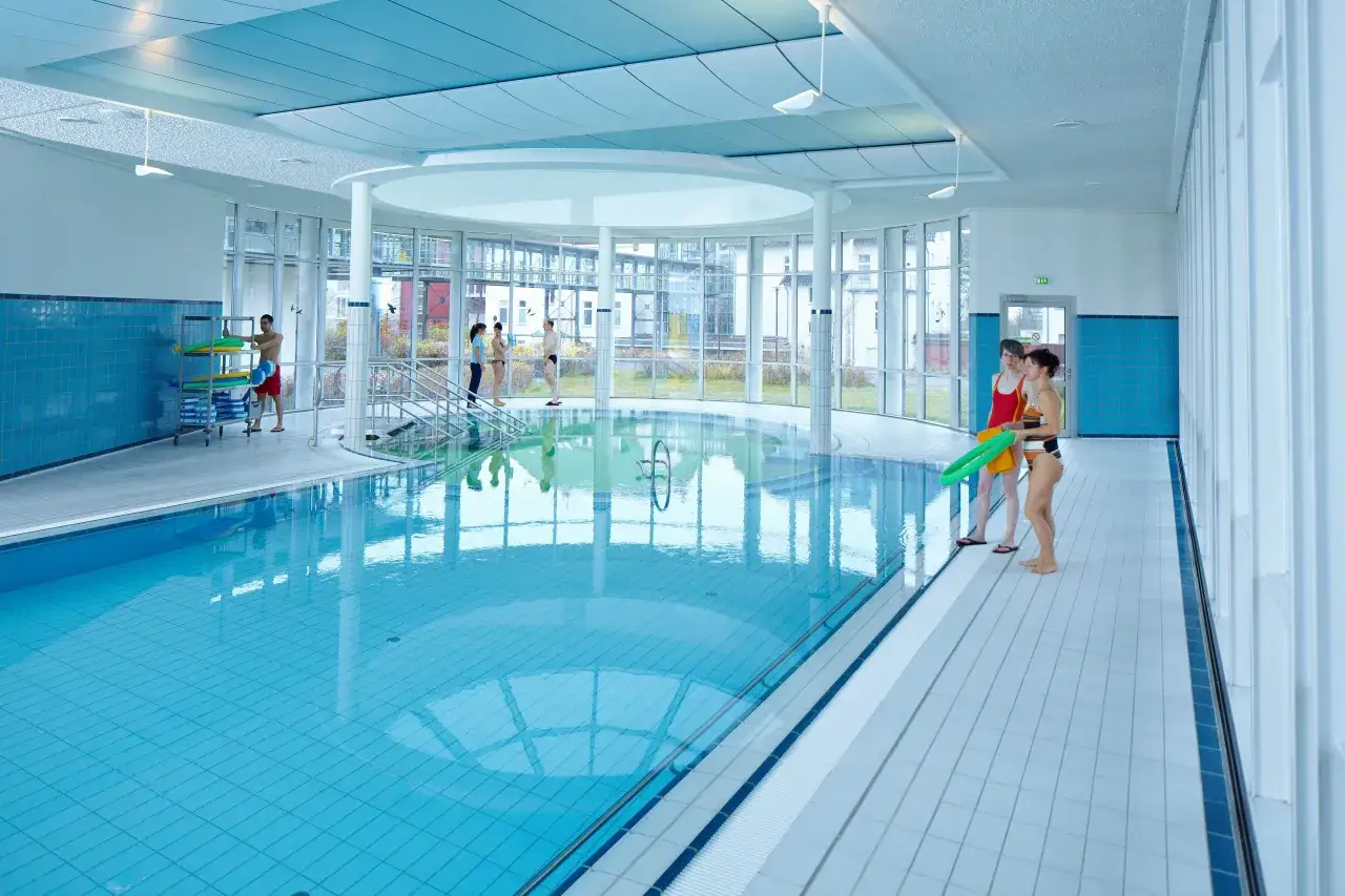 Menschen in Badekleidung am Beckenrand eines Hallenbades mit türkisfarbenem Wasser.