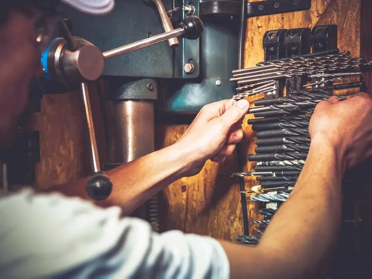 Ręce wybierające wiertło z uchwytu obok wiertarki stołowej.