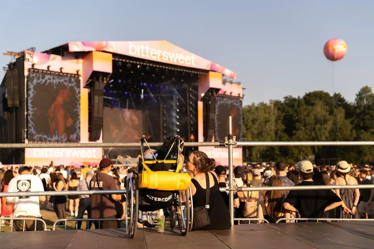 Dwie osoby, jedna na wózku, dzielą intymny moment na festiwalu bittersweet. W tle scena i tłum.