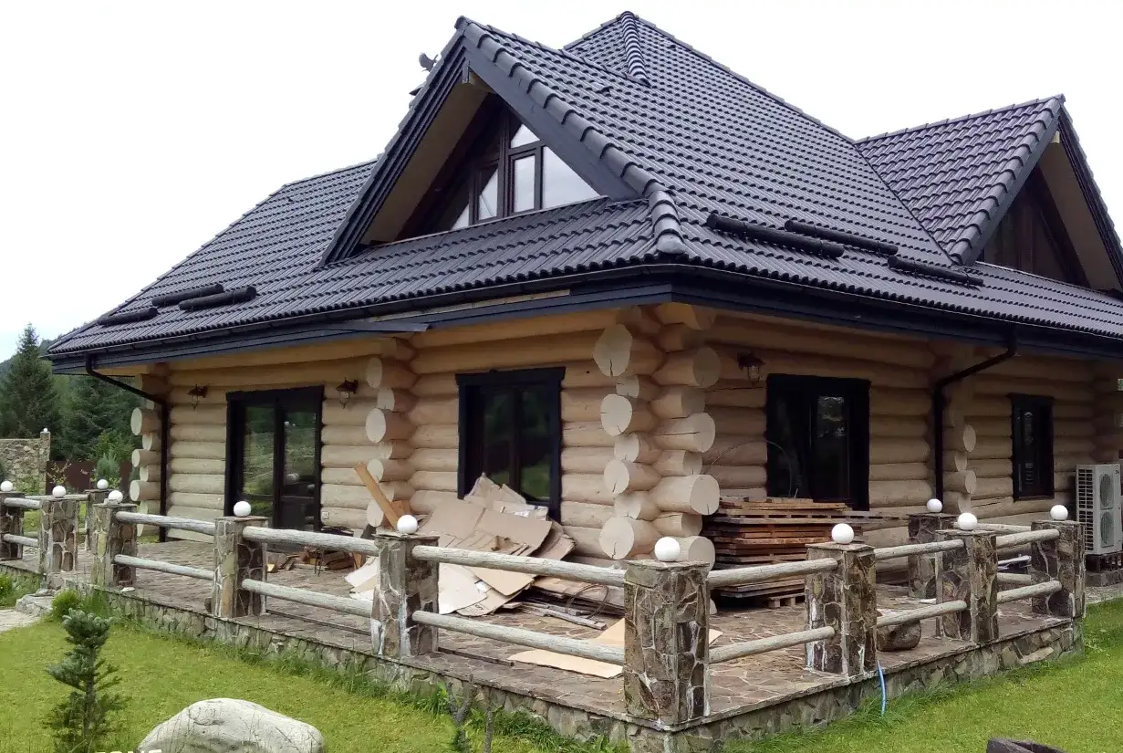 Drewniany dom z bali z czarnym dachem i tarasem, otoczony zielenią.