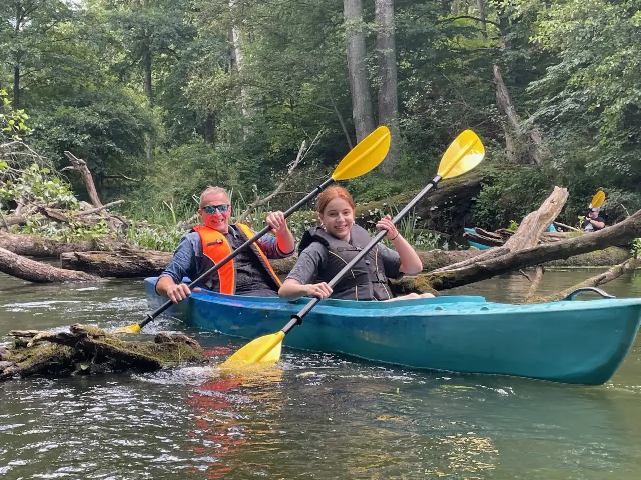 Dwie osoby w kajaku na zalesionej rzece, wiosłujące wśród powalonych drzew.