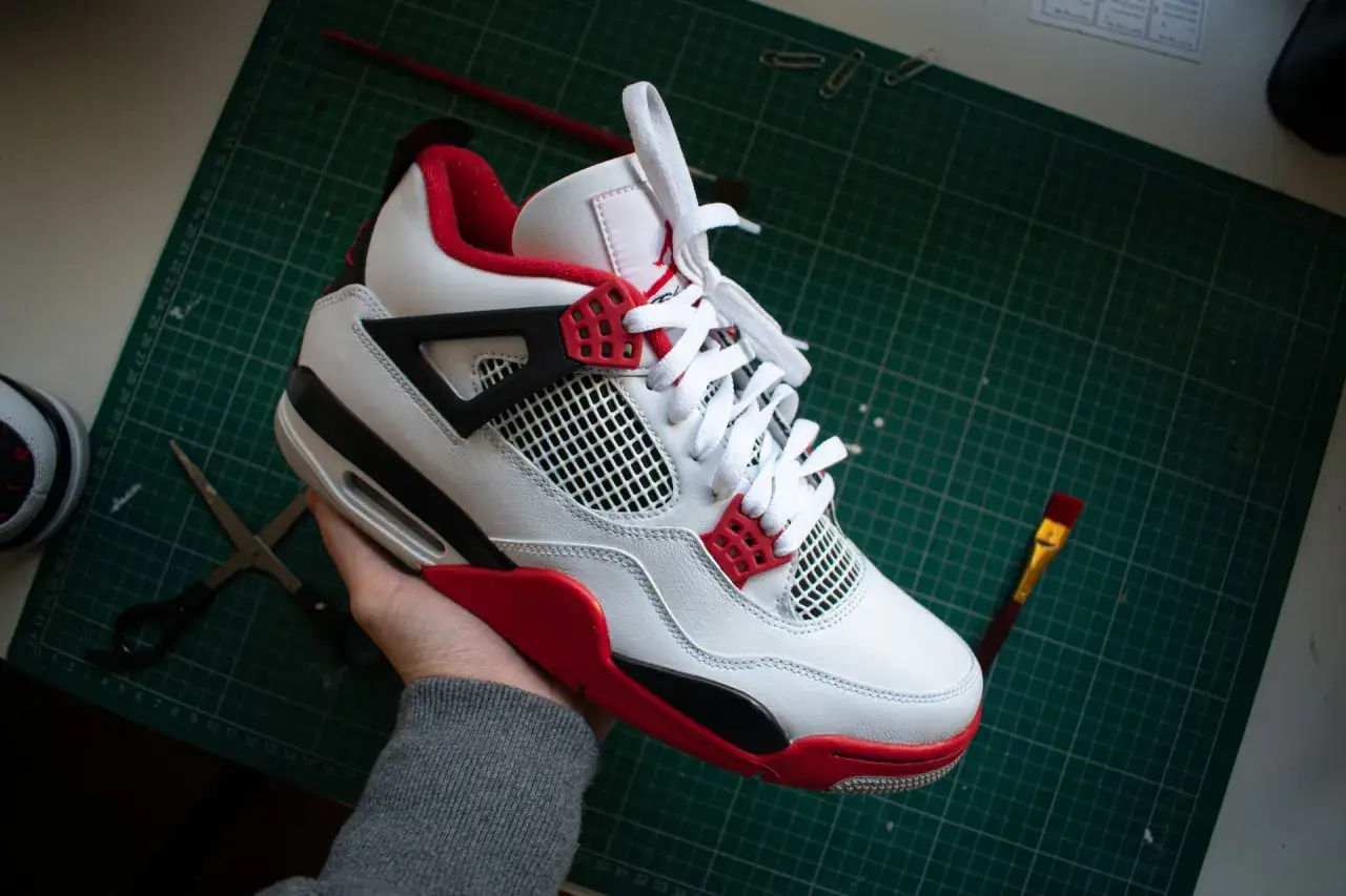Białe buty Jordan 4 z czerwonymi akcentami, które wyglądają jak podróbki. Na stole leżą nożyczki i pędzel.