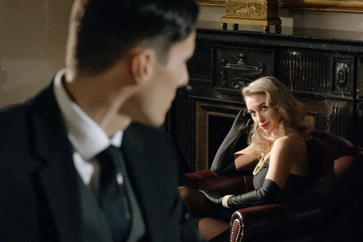 Elegancka kobieta w czarnej sukni i rękawiczkach, z perłami na szyi, wpatruje się w mężczyznę. Scena pełna nls uwodzenia.
