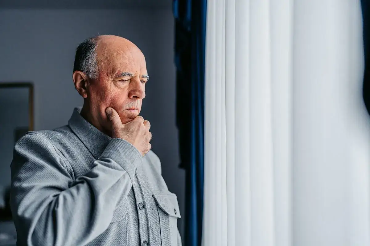 Starszy mężczyzna w szarej koszuli zamyślony przy oknie.