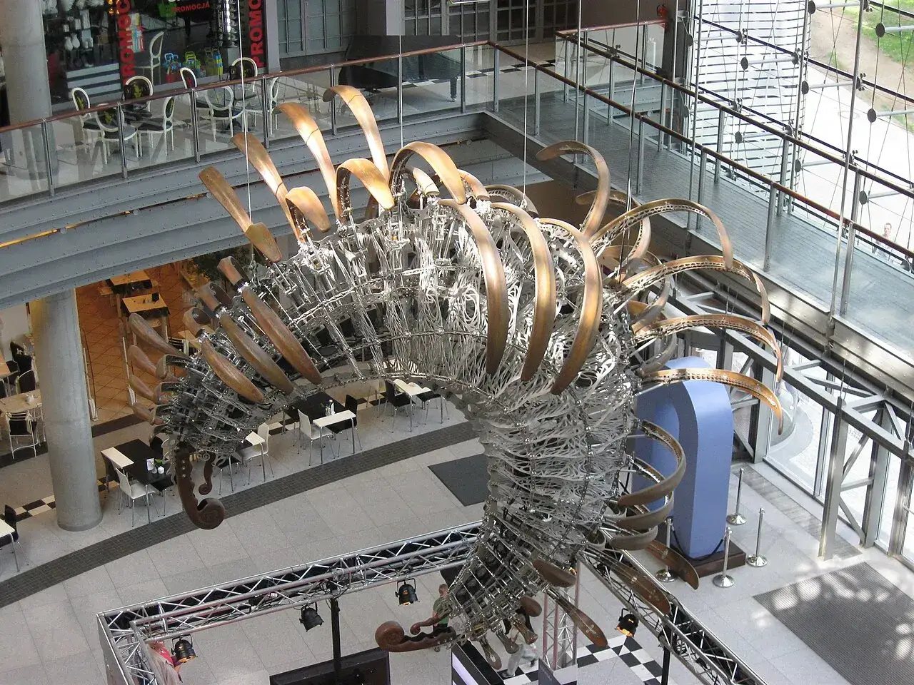Duża, metalowa rzeźba przypominająca szkielet dinozaura w centrum handlowym.