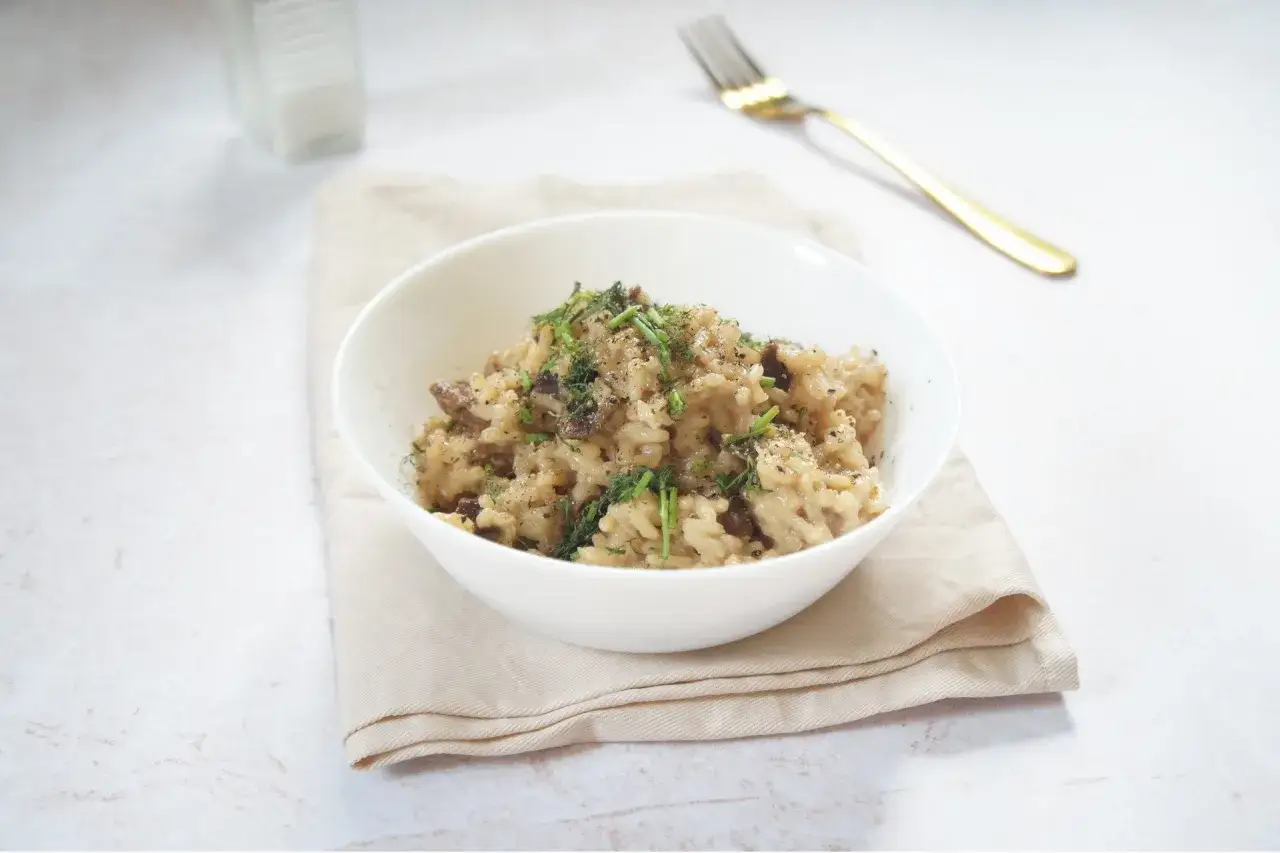 Miseczka kremowego risotto z grzybami, posypana ziołami i pieprzem, obok złoty widelec.
