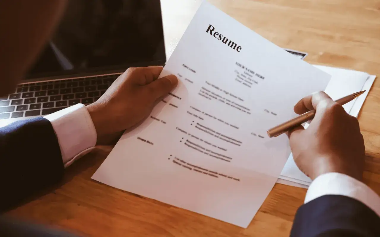 Dłoń trzymająca długopis nad dokumentem CV obok laptopa.