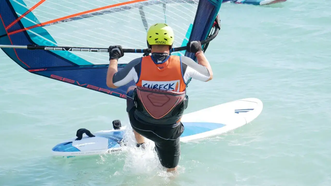Osoba w kasku i kamizelce uprawia windsurfing na turkusowej wodzie.