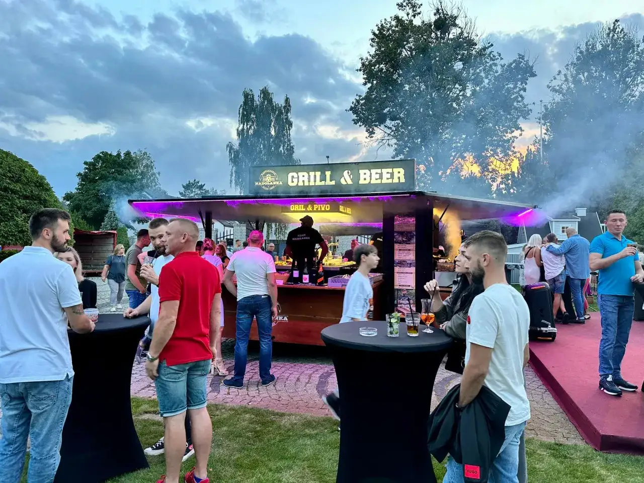 Ludzie przy stoisku z grillem i piwem, dym i fioletowe światła.
