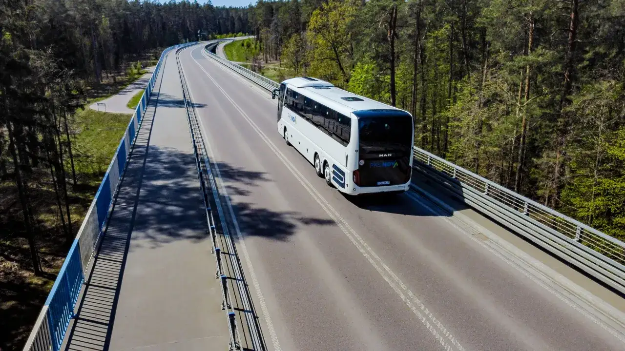 Biały autobus PKS Nova jedzie po moście wśród drzew.