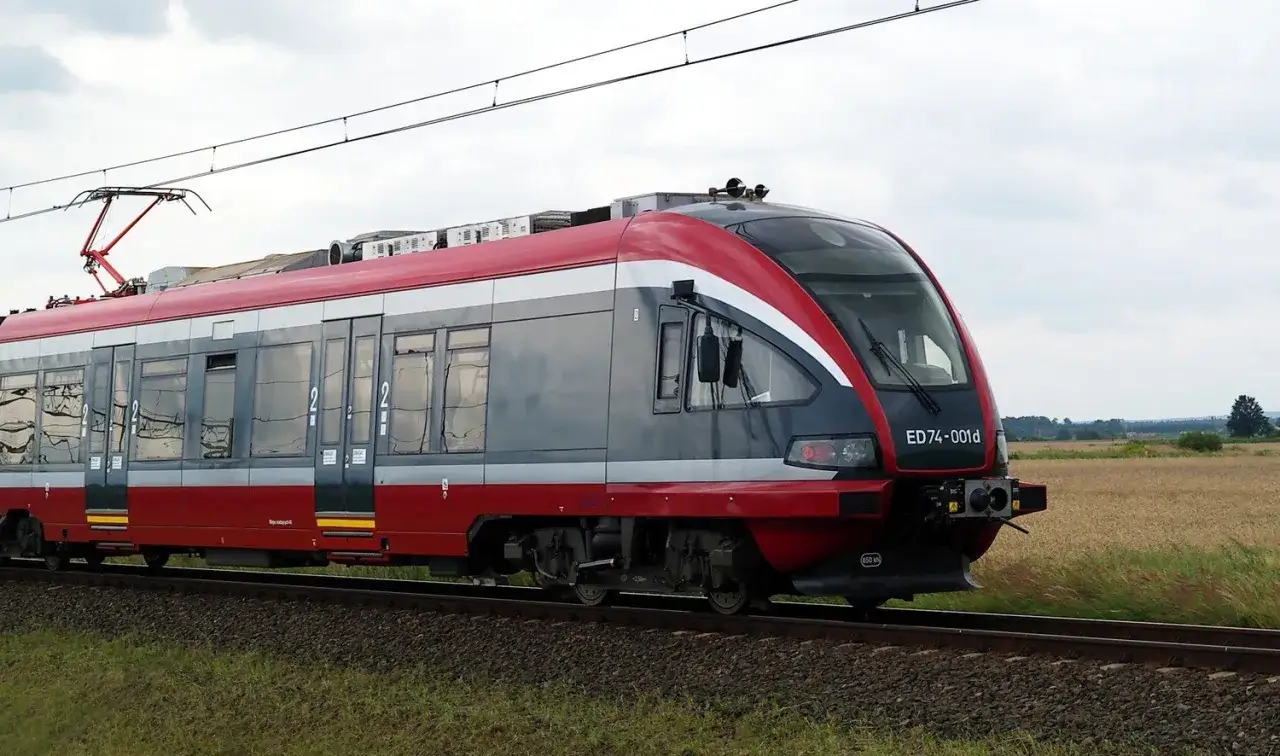 Czerwono-szary pociąg elektryczny ED74-001d na torach, w tle pole i niebo.