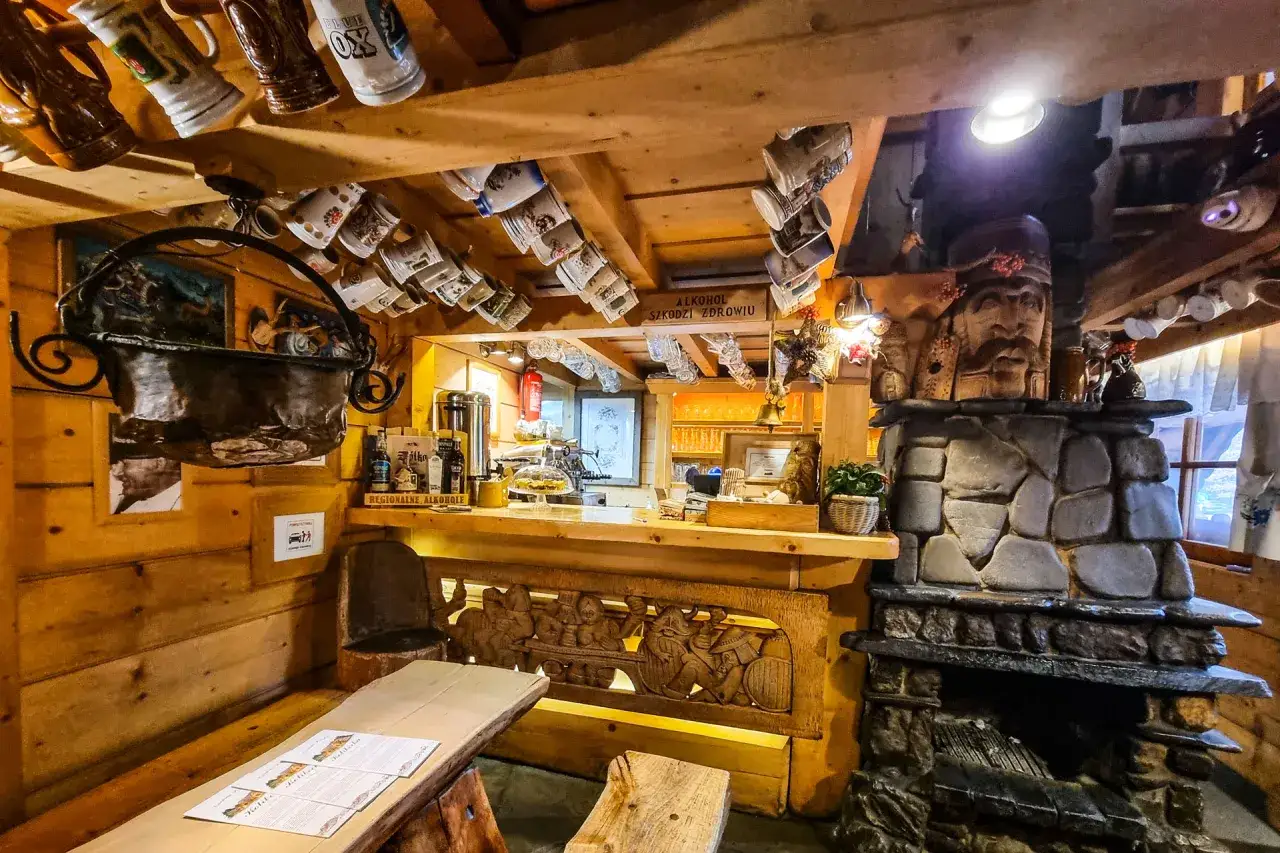 Drewniane wnętrze karczmy z kamiennym kominkiem, rzeźbionym barem i wiszącymi kubkami.