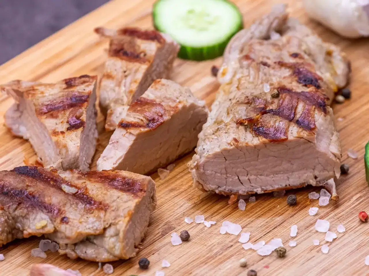 Soczyste polędwiczki wieprzowe i karczek z grilla, pokrojone na desce z drewnianymi ziarnami, posypane solą i pieprzem.