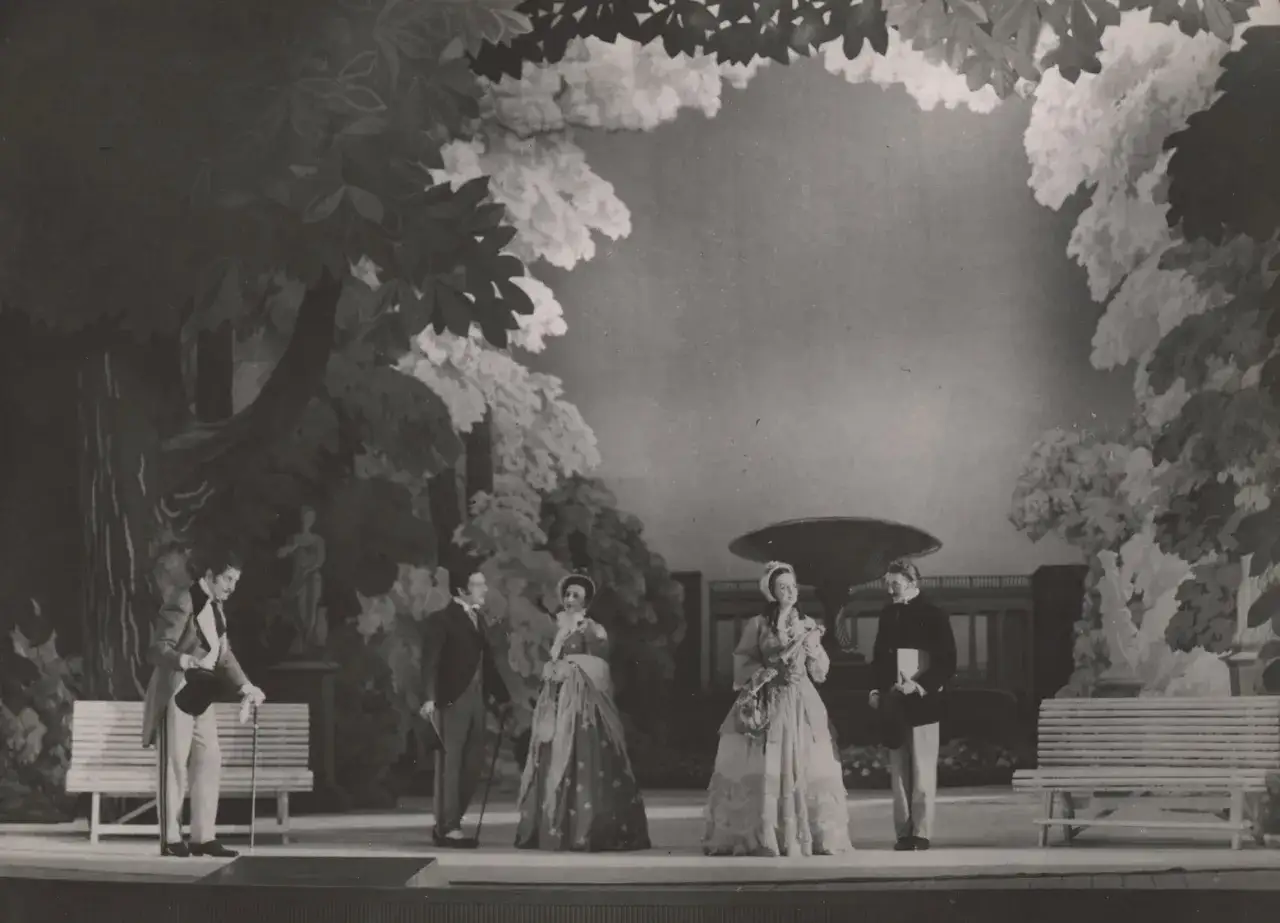 Scena z przedstawienia teatralnego, gdzie aktorzy w strojach z epoki odgrywają scenę w ogrodzie. Reżyser teatralny Szyfman zadbał o szczegóły scenografii.
