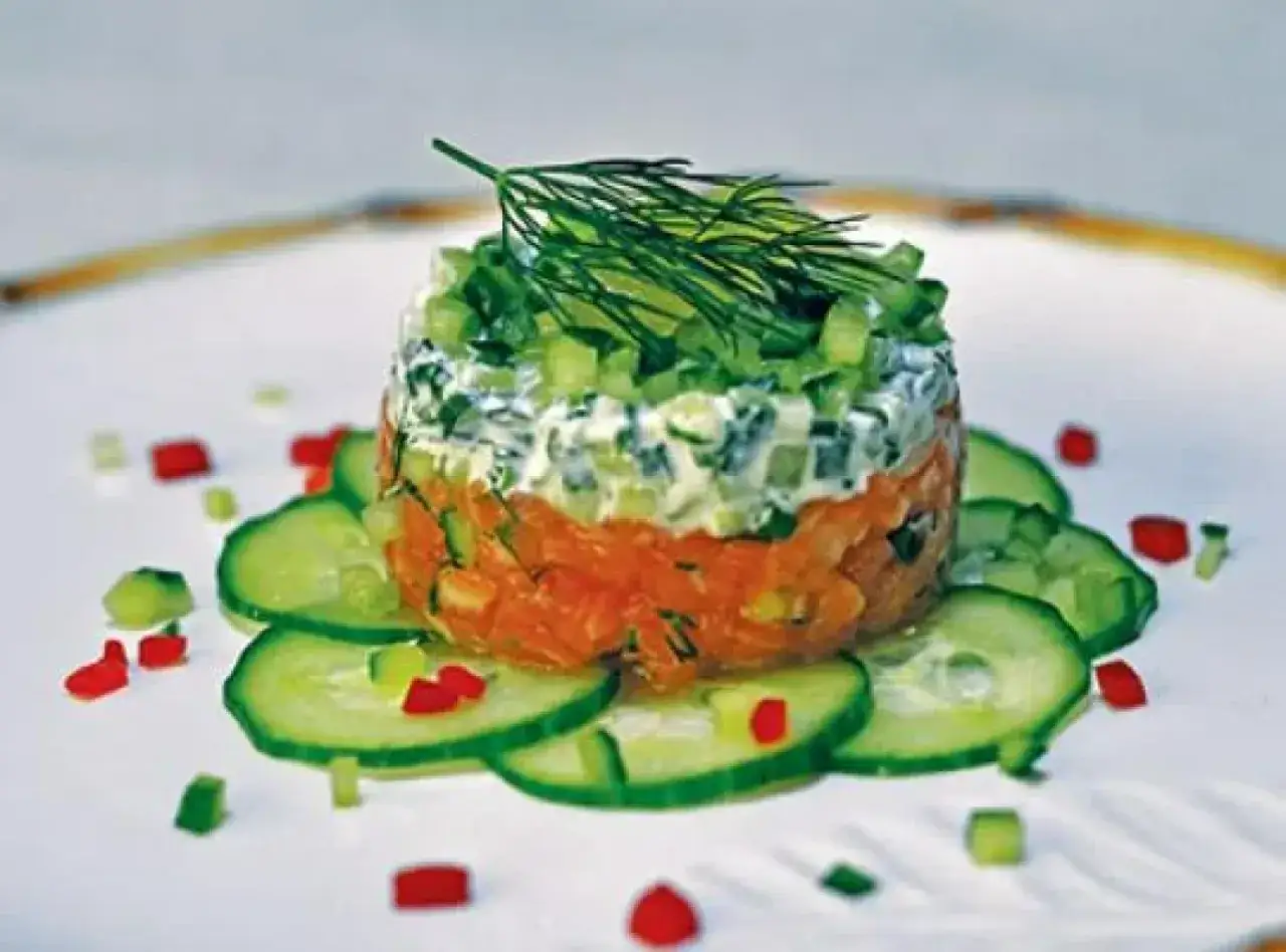 Wykwintny tatar z łososia, ułożony warstwowo z kremowym sosem i świeżymi warzywami, ozdobiony koperkiem i plasterkami ogórka.
