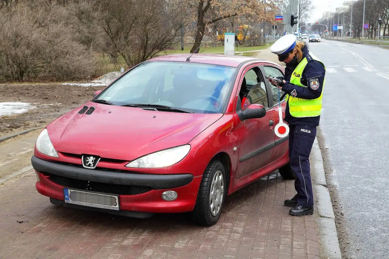 Policjantka w kamizelce odblaskowej i czapce spisuje kierowcę przy czerwonym samochodzie.