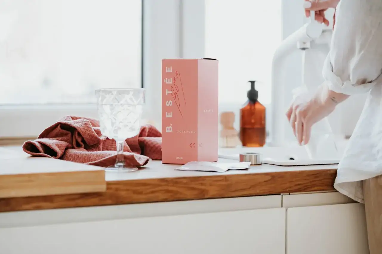 Różowe pudełko z kolagenem B. ESTÉE, szklanka wody i ręcznik. To estetyczne zdjecia produktowe.