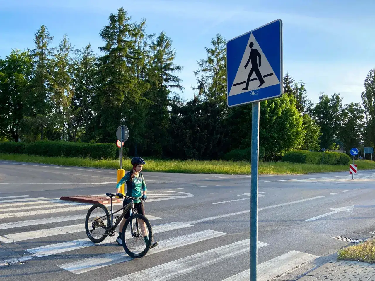 Rowerzystka w kasku prowadzi rower po pasach dla pieszych obok znaku drogowego.