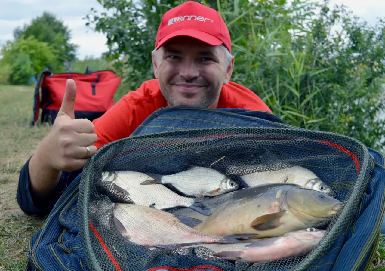Szczęśliwy wędkarz z okazałym połowem ryb w podbieraku. W tle czerwony plecak i zielona roślinność. Gotowa zanęta do metody zadziałała!