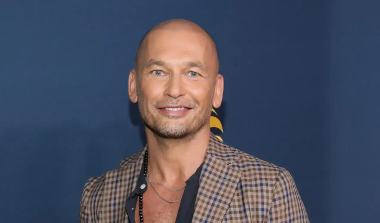 Tomasz Wygoda, łysy mężczyzna z niebieskimi oczami, uśmiecha się, ubrany w kratowaną marynarkę i naszyjnik.