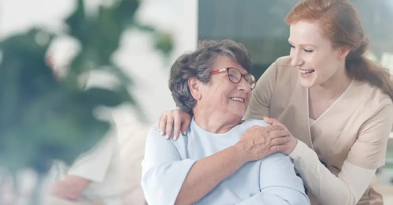 Uśmiechnięta opiekunka obejmuje starszą panią. Pytanie "ile bierze opiekunka do osoby starszej" nabiera sensu, gdy widzimy taką troskę.