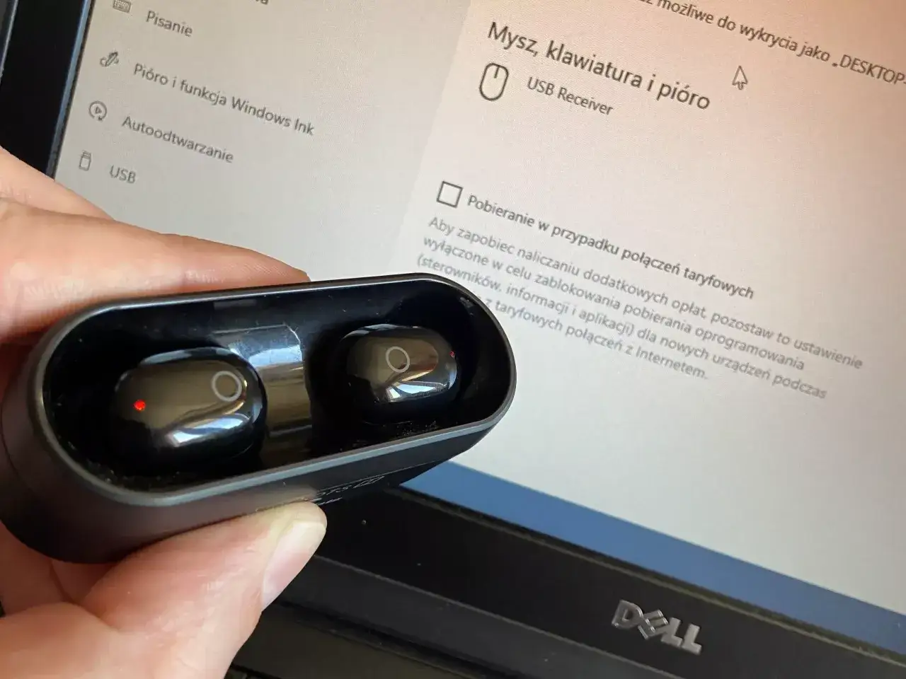 Dłoń trzyma czarne słuchawki w etui, obok ekranu laptopa z ustawieniami, pokazując jak sparować słuchawki z laptopem.