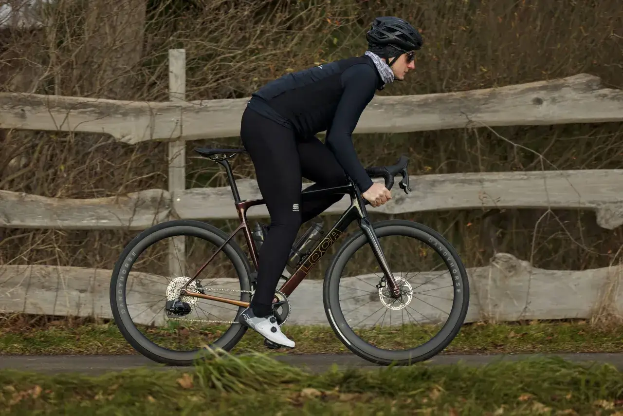 Kolarz w czarnym stroju pędzi na rowerze road plus obok drewnianego płotu.
