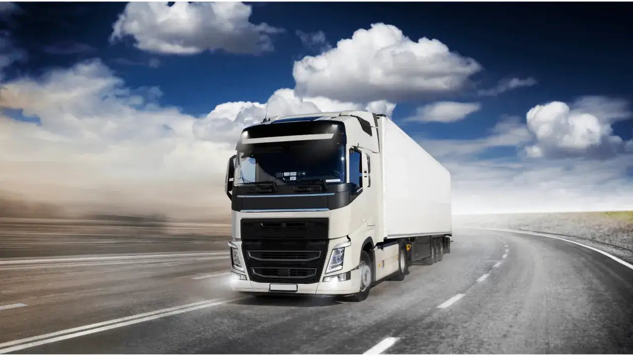 Biała ciężarówka z naczepą jedzie po pustej drodze pod błękitnym niebem z chmurami.