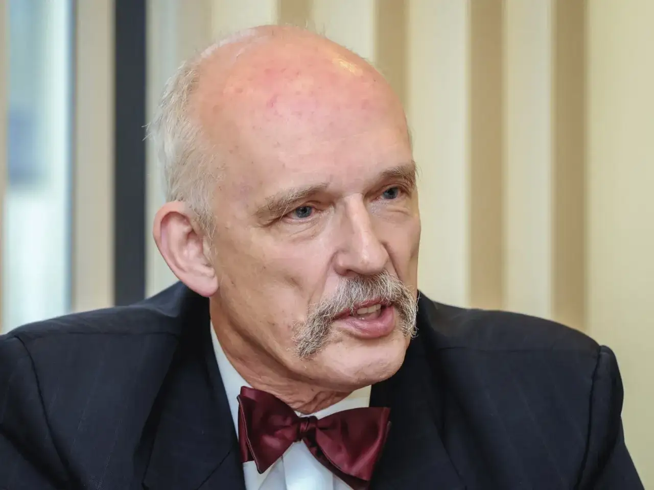 Janusz Korwin-Mikke w garniturze i bordowej muszce, z charakterystycznymi wąsami.