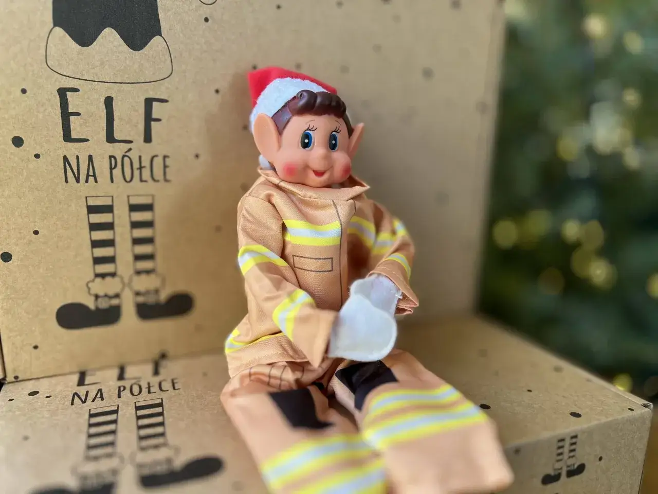Uroczy elf na półce w stroju strażaka siedzi na kartonie z rysunkami elfich nóg.