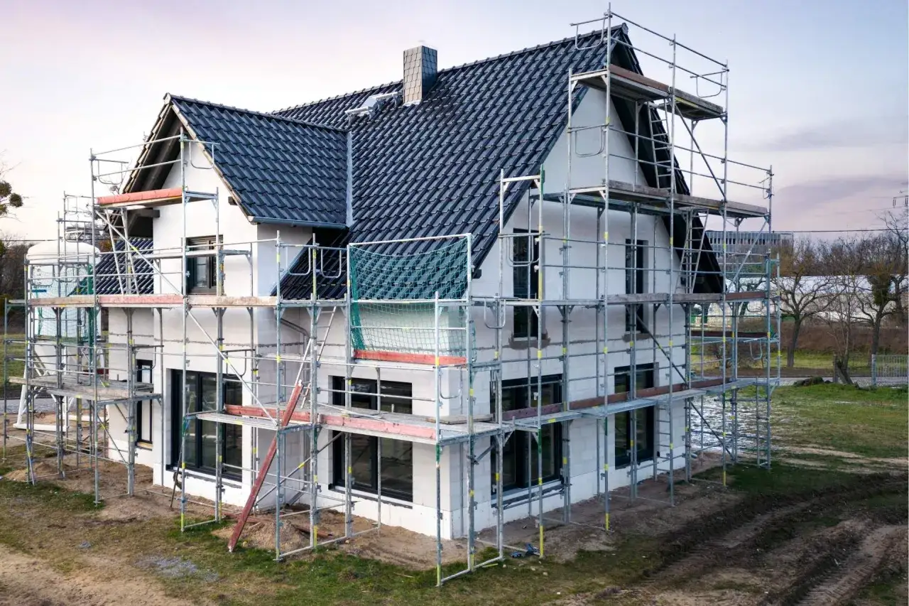 Dom w budowie, otoczony rusztowaniami, z czarnym dachem i białymi ścianami. To pierwszy etap, jak zbudować dom.