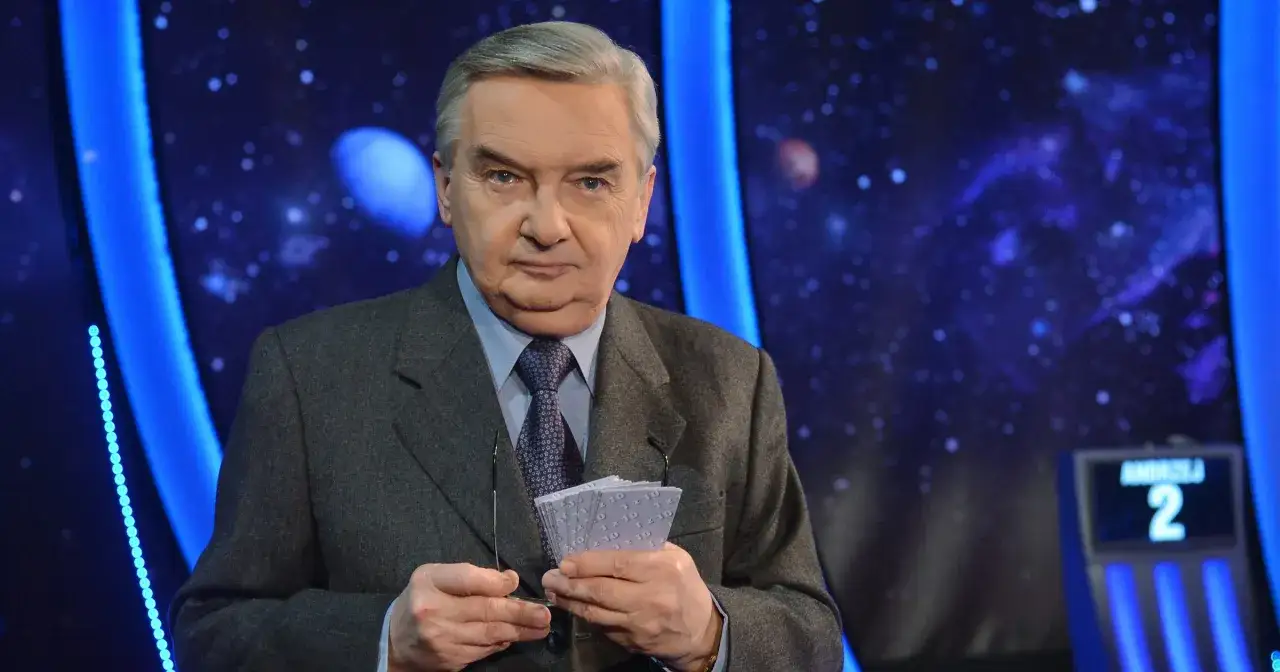 Starsza osoba w garniturze trzyma karty z napisem "1 z 10", gotowa do emisji.