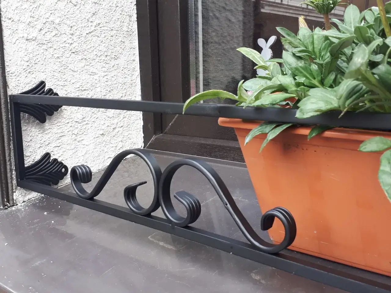 Elegancka barierka na parapet bez wiercenia, ozdobiona kutymi elementami, chroni doniczkę z zielonymi roślinami.