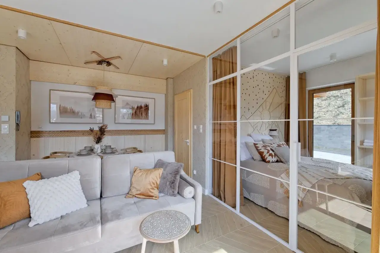 Przytulne pokoje i apartamenty Wierchy Zakopane. Salon z sofą i widokiem na sypialnię z łóżkiem, w góralskim stylu.