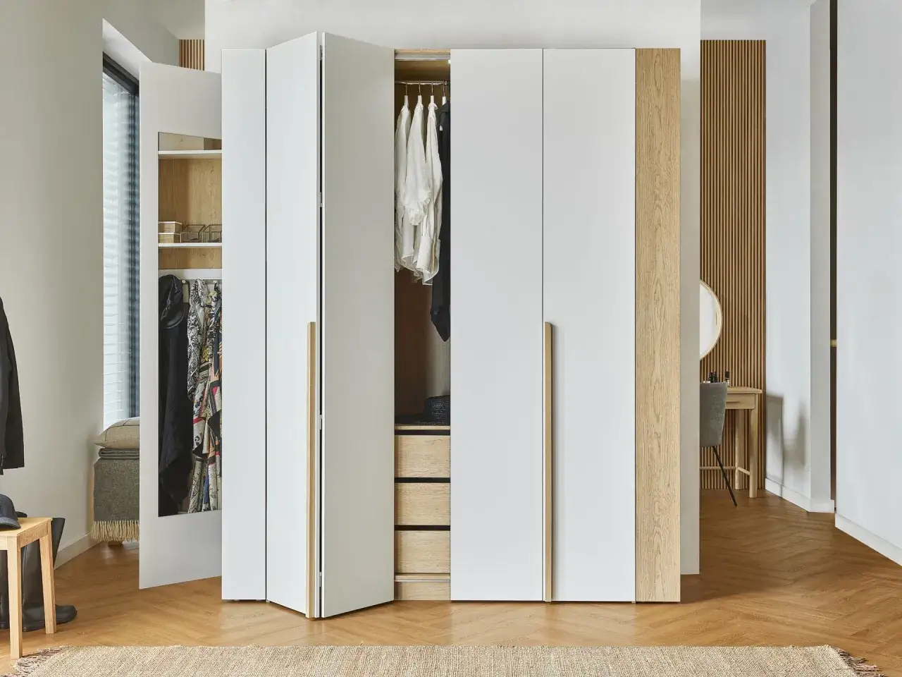 Nowoczesna garderoba w sypialni za ścianką, z białymi frontami i drewnianymi akcentami. W środku wieszaki z ubraniami i szuflady.