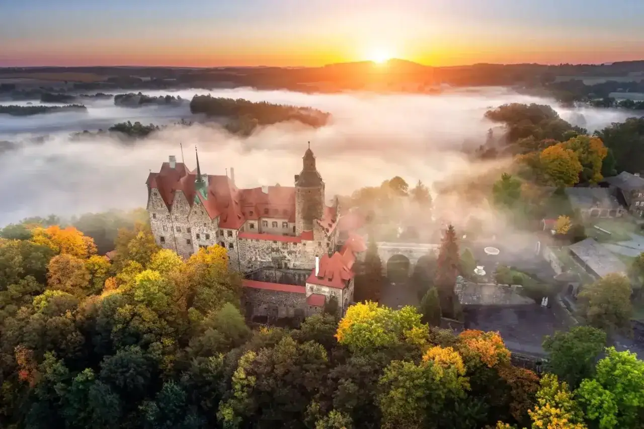 Zamek Książ otoczony jesiennymi drzewami, wyłaniający się z porannej mgły. Słońce wschodzi nad wzgórzami, malując niebo na pomarańczowo.