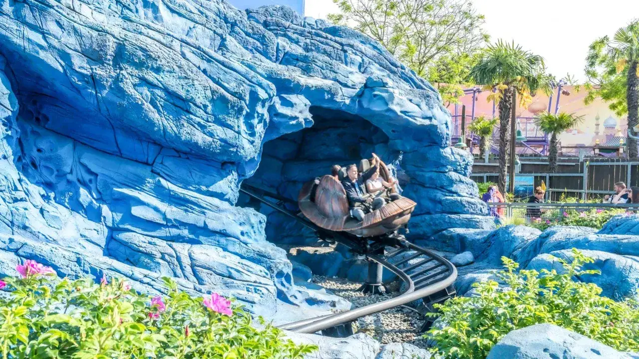 Personas en un coche de montaña rusa saliendo de una cueva azul rocosa, con palmeras y un paisaje de cuento de hadas al fondo.
