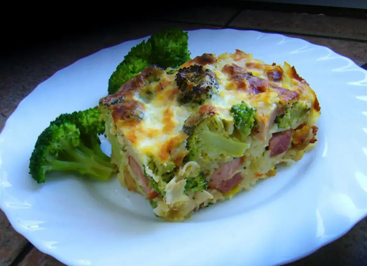 Kawałek zapiekanki makaronowej z brokułami i szynką, podany na białym talerzu z dodatkiem różyczek brokułu.