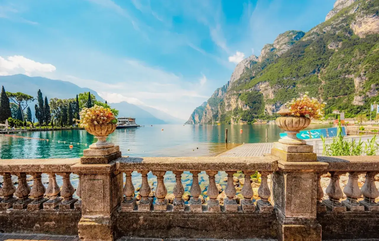 Kamienna balustrada z wazonami kwiatów prowadzi do widoku na turkusowe **Jezioro Garda** i góry. Statek kołysze się na wodzie, a niebo jest błękitne.