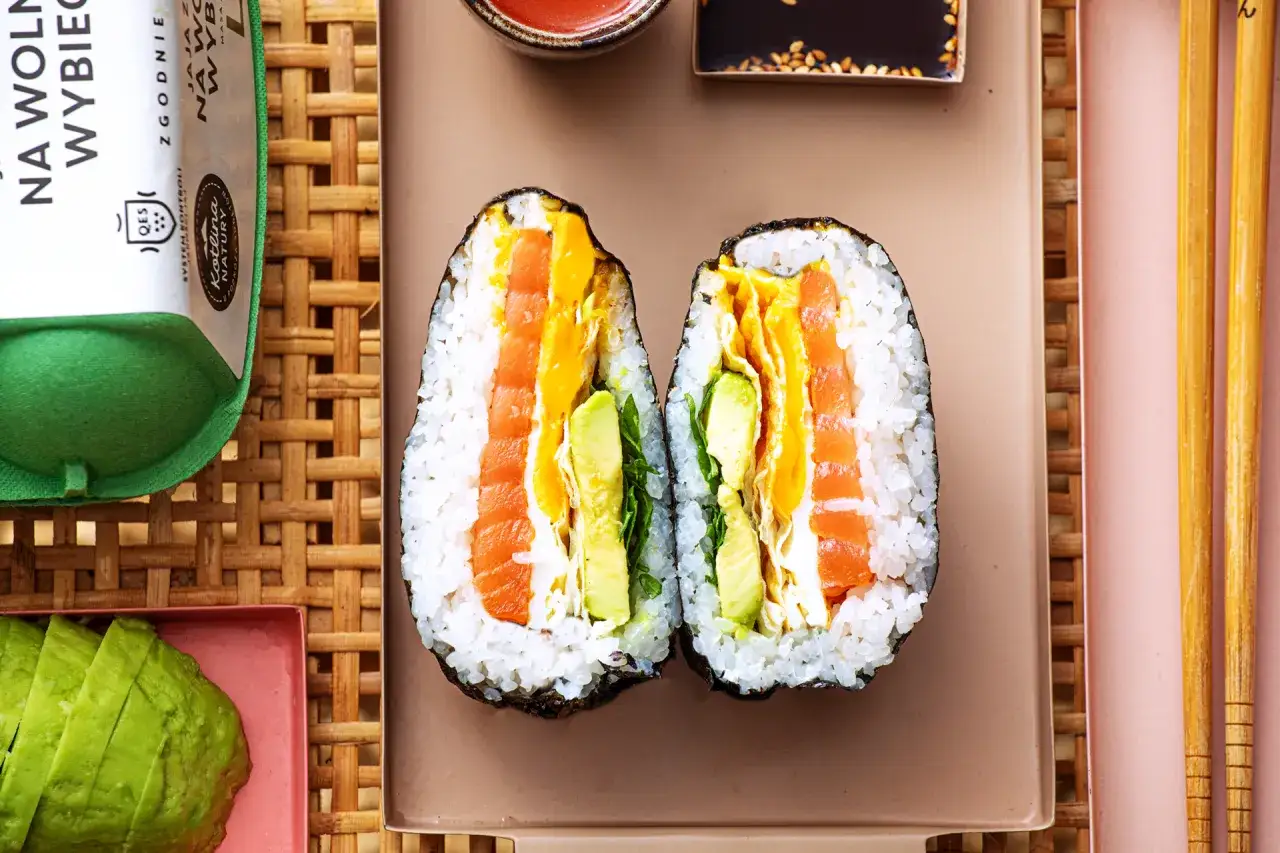 Dwa połówki onigiri z łososiem, awokado i jajkiem, podane na talerzu z pałeczkami.