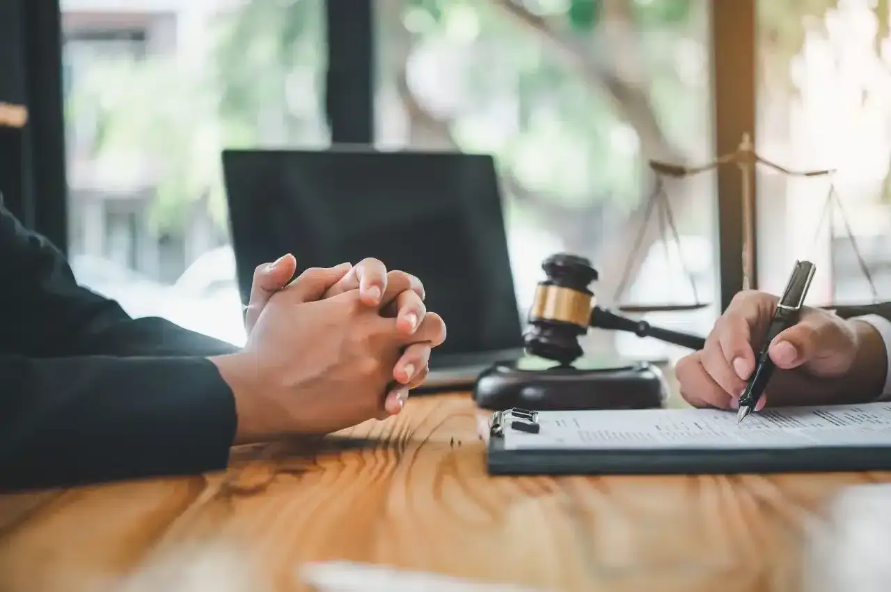 Dłonie splecione na stole, młotek sędziowski, waga sprawiedliwości i ręka pisząca na dokumencie.