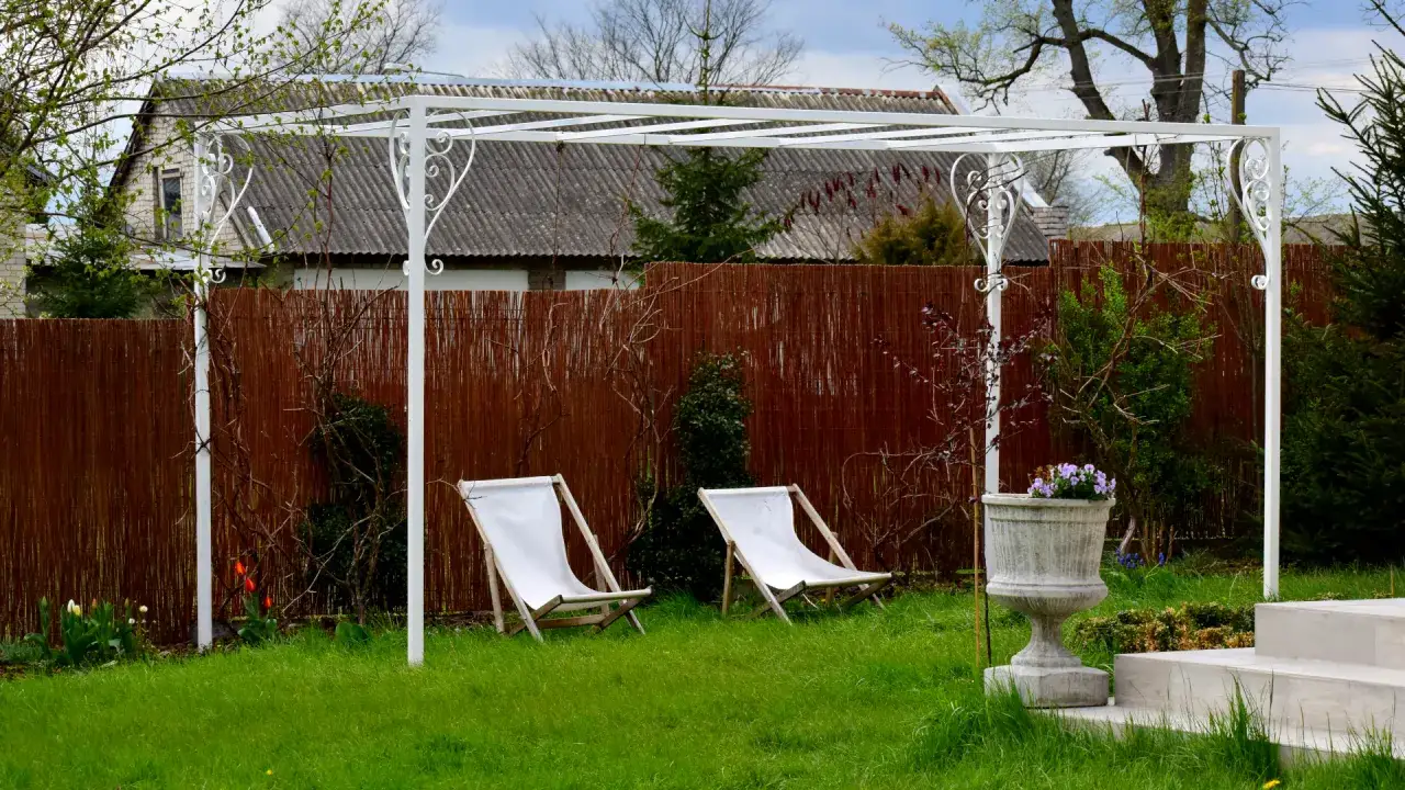 Biała pergola ogrodowa z dwoma leżakami i donicą z kwiatami.