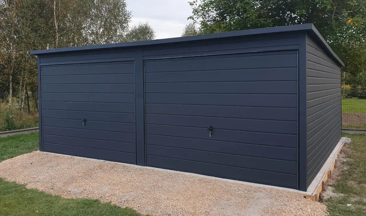 Nowoczesny garaż z płaskim dachem, z dwoma ciemnoszarymi bramami, otoczony zielenią i żwirem.