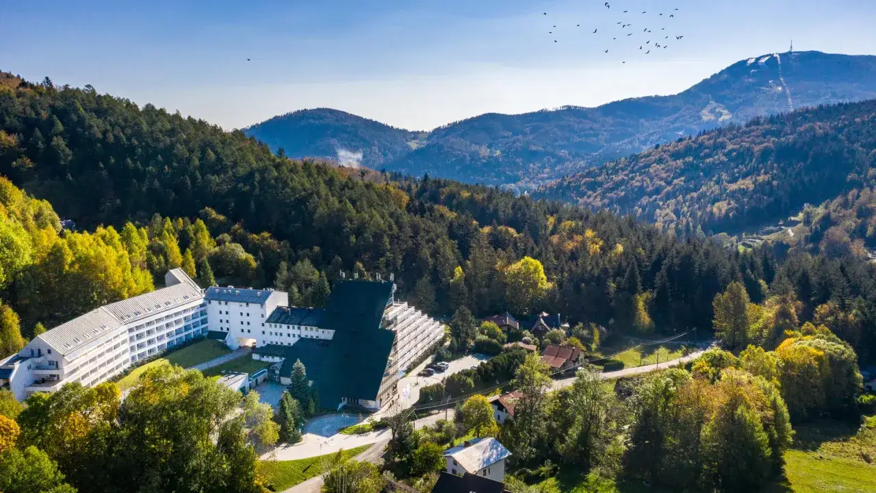 Malowniczy hotel przy stoku narciarskim w Polsce, otoczony jesiennym lasem. W oddali góry i niebo z ptakami.
