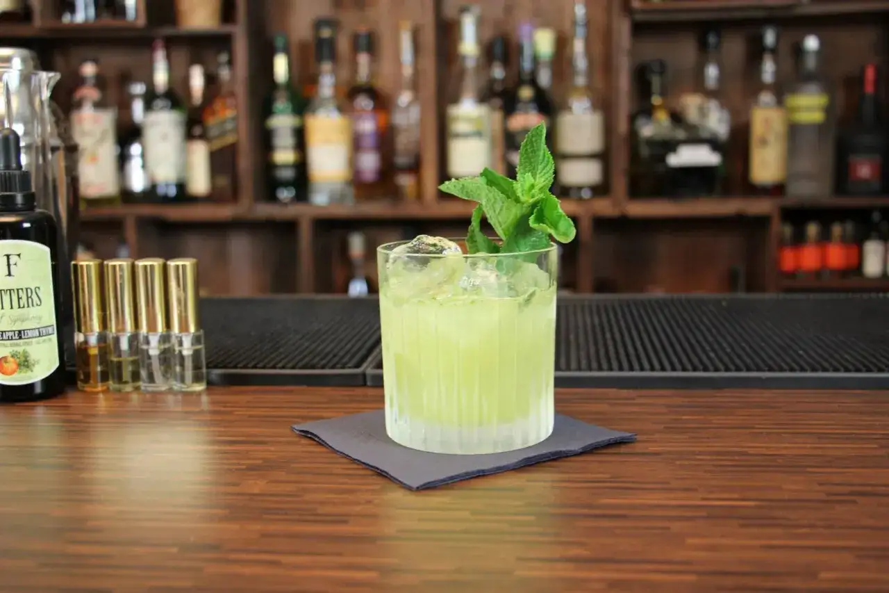 Ein grüner Tequila-Cocktail mit Minze und Eis auf einem Holztresen, im Hintergrund Regale mit Flaschen.