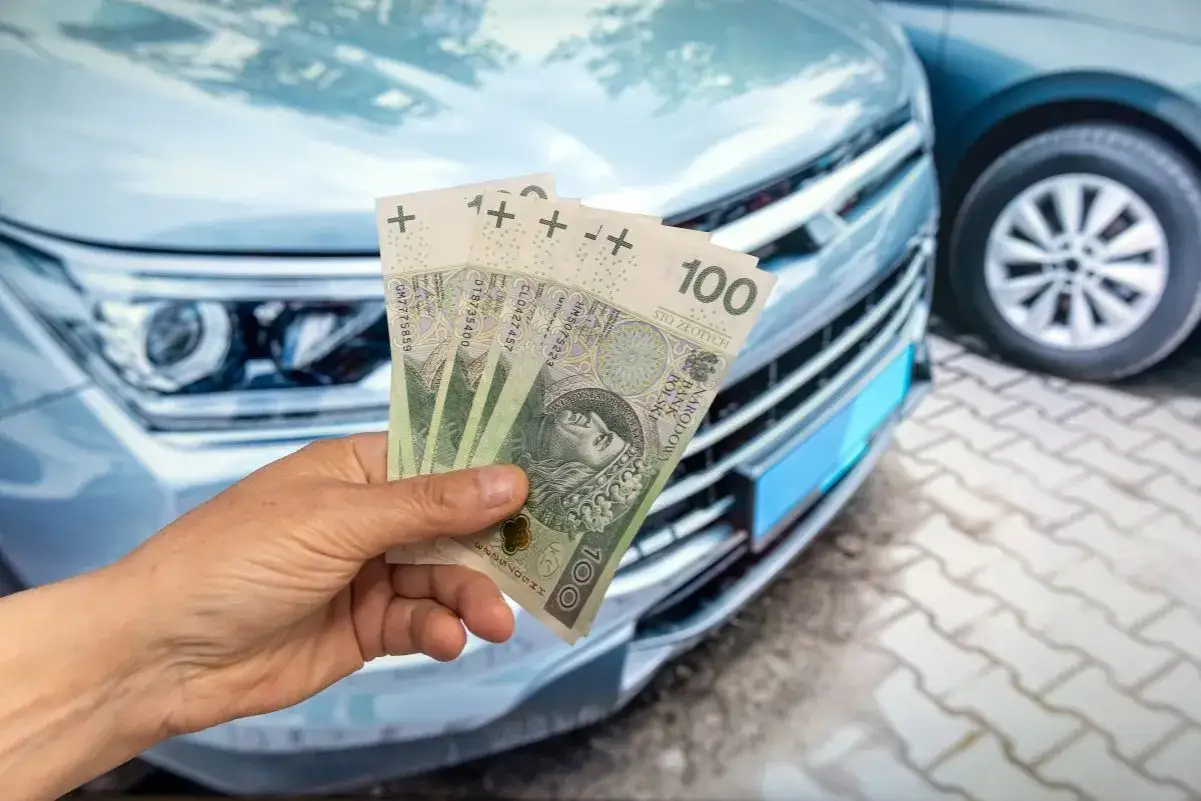 Dłoń trzyma polskie banknoty na tle przodu samochodu.