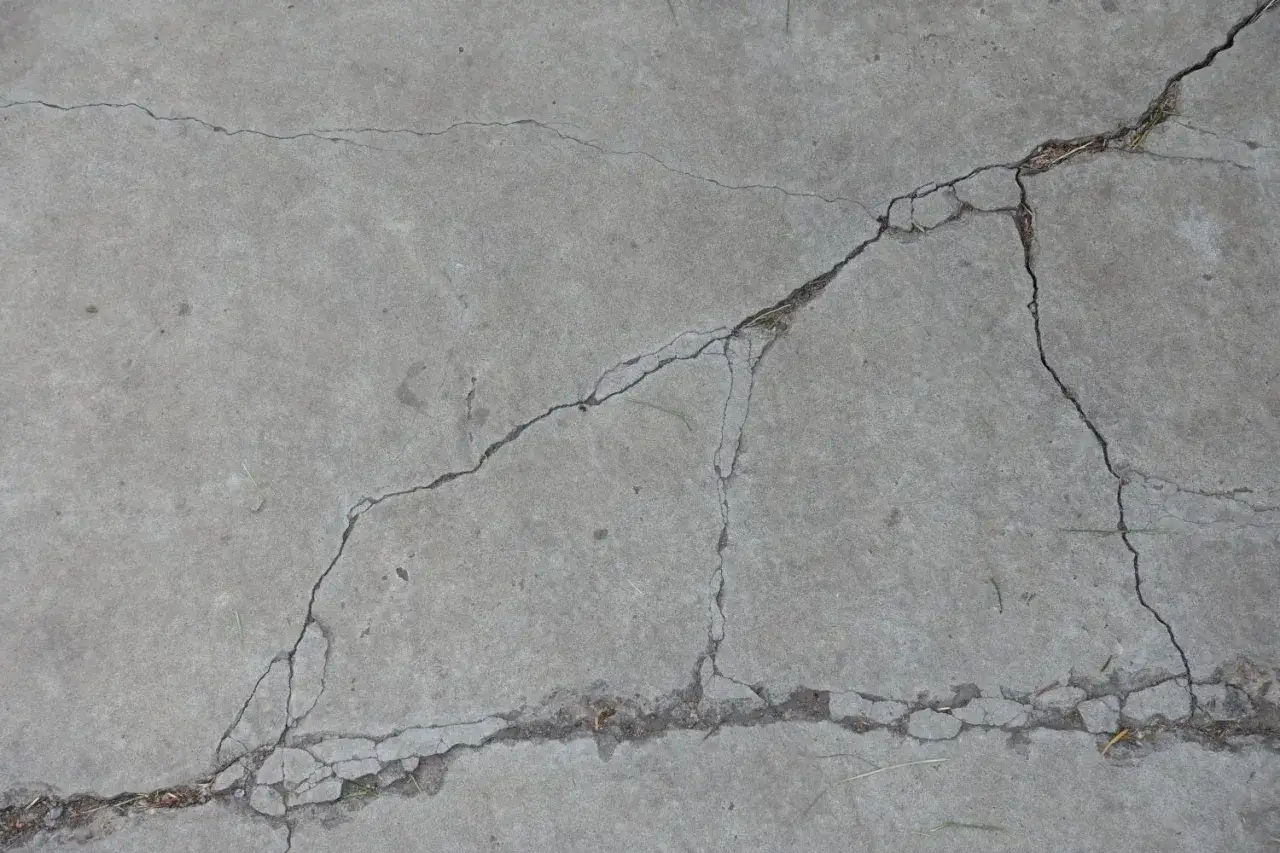Popękany beton, jakby po długim okresie podlewania, z widocznymi szczelinami i drobnymi źdźbłami trawy.