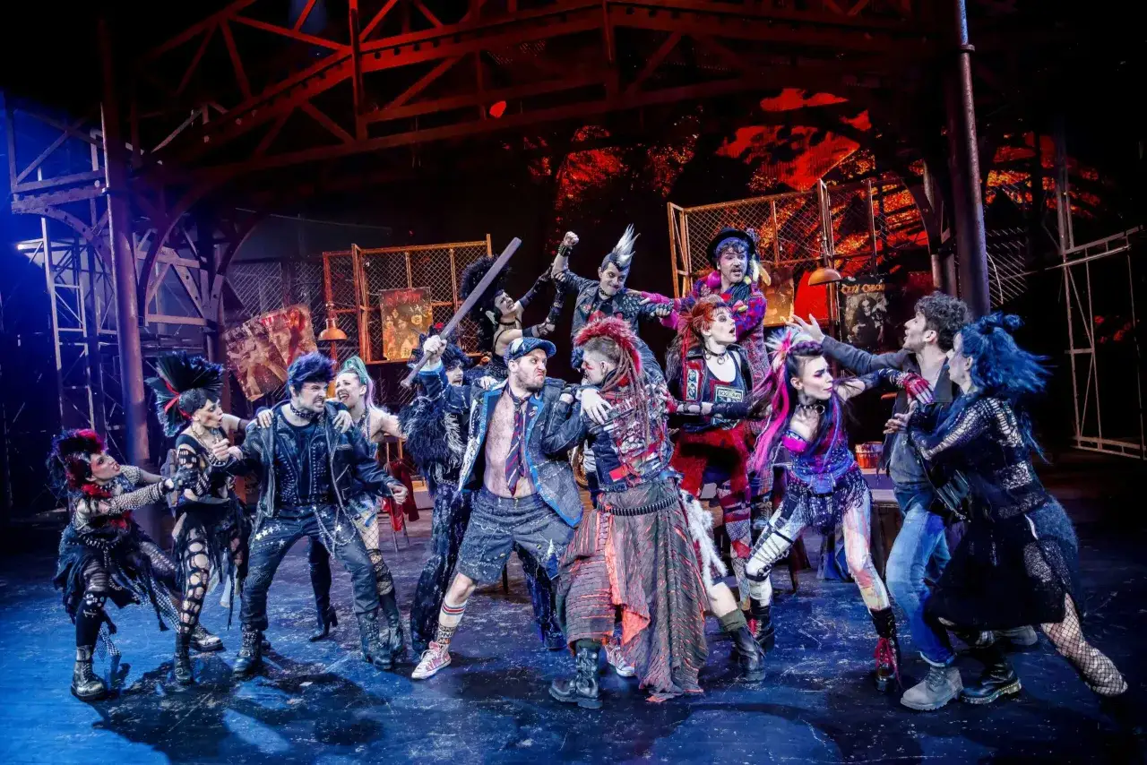 Grupa punkowych postaci na scenie, w tym z irokezami i w skórzanych kurtkach, w dynamicznej scenie z musicalu "We Will Rock You".