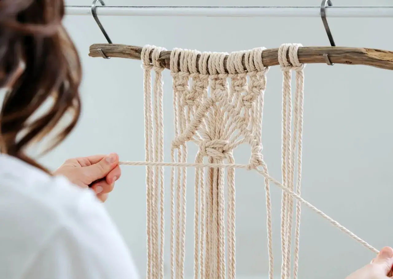 Ręcznie robiona makrama zamiast firanki, tworzona przez kobietę. Delikatne sznurki oplatają drewniany patyk, tworząc piękny wzór.