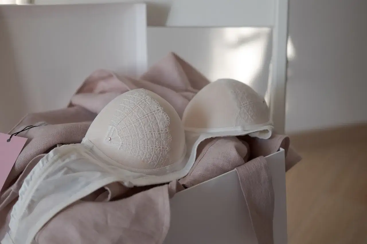 Dwa białe biustonosze z koronkowymi zdobieniami, ułożone na pudrowej tkaninie.